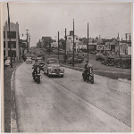 Two policeman on motorcycles escort cars eastbound on East 12th Street at 14th Avenue in the Clinton Park district of Oakland, California