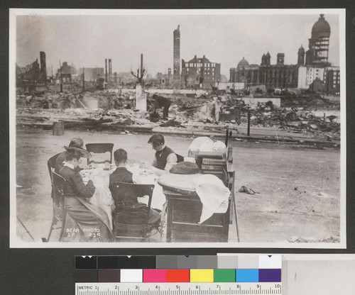 [Family at table in street among ruins. City Hall in background.]