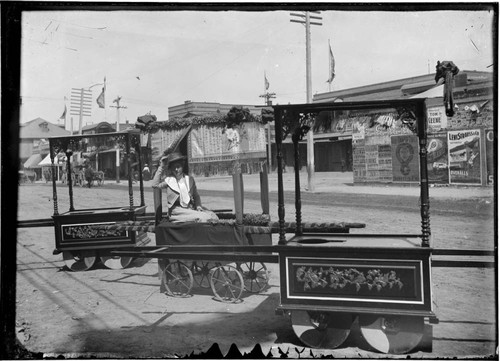 White woman posing on ceremonial handcart, with street and advertisements in background