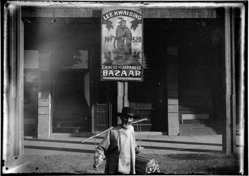 Man carrying bundles, under advertisement for "Lee Kwai Sing, No. 520, Chinese and Japanese Bazaar"