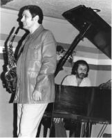 Art Pepper holding a saxophone, with Milcho Leviev in the background [descriptive]