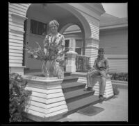 Mertie West stands next to a vase of flowers while Richard Siemsen sits on the porch behind her, Los Angeles, 1943