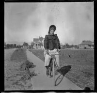 Earl Kellum, dressed in women's attire, rides a bicycle along a sidewalk, Los Angeles, about 1899