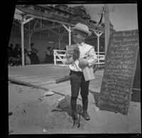 Lester Shaw stands by a chalkboard sign holding firecrackers, Santa Monica, 1901