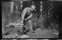 Sheep herder traveling with H. H. West and company removing the coals from a Dutch oven, Shasta County, 1917