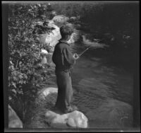 H. H. West, Jr. fishes in Matilija Creek while standing on a small rock, Ojai vicinity, about 1925