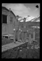 Two uniformed servicemen pose on the walkway in front of their quarters, Dutch Harbor, 1943