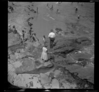 Mertie West and H. H. West, Jr. are viewed from above as they walk over boulders towards the beach in La Jolla, San Diego, about 1930