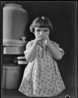 Photograph of a little girl drinking a glass of water, standing in front of a stoneware Arrowhead water cooler, Los Angeles, 1935