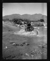 Man crossing a stream on a motorcycle, circa 1920
