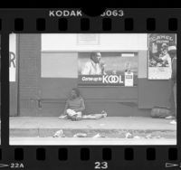 Man sitting on skid row sidewalk below cigarette ads in Los Angeles, Calif., 1986