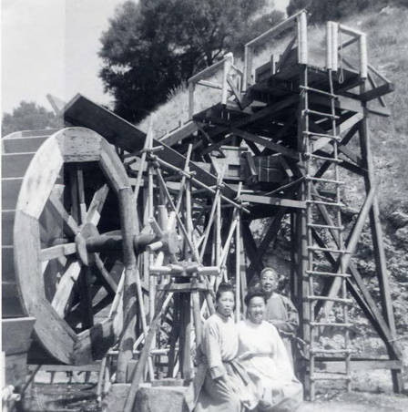 Two women and a man sitting in front of a water wheel. On the back of the photo it reads "Slosson Jong- Lost Horizon 1972"