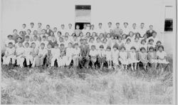 Large group of Japanese school children and teachers seated and standing outside next to a building