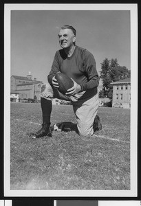 University of Southern California assistant football coach Sam Barry, in dark sweatshirt, kneeling and holding football, on Bovard Field, 1947