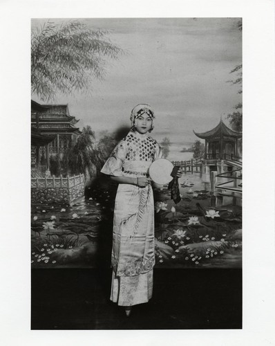 Actress in classical dress and lotus feet holds a round fan on background of Chinese pavilions over a lotus pond /