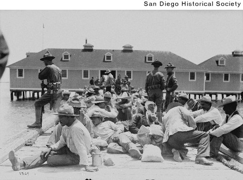 Mexican refugees sitting on a dock near a boathouse under the guard of US Soldiers