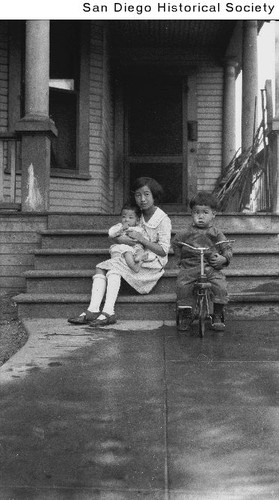 Pearl Hom sitting on the steps of the Tom Sun Mow residence holding William while Gene sits on a tricycle