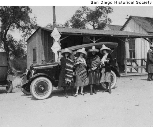 Four women wearing sombreros and blankets standing in front of an automobile