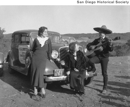 Two women sitting on the front of an automobile while being serenaded by a Mexican singer