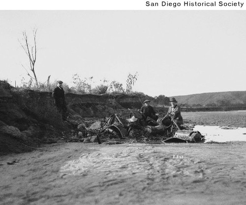 Three men in a river bed with a half buried Ford automobile following a ...