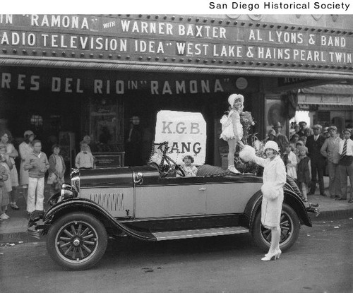 A young girl standing on the back of a 1928 Durant automobile parked in front of a movie theater