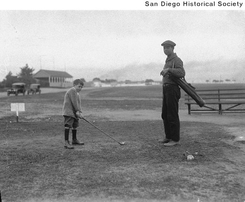 A small boy playing golf with an adult holding the golf bag