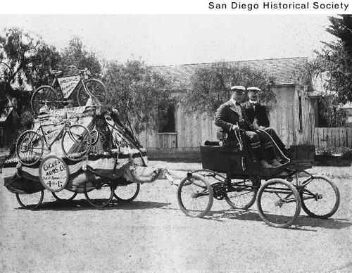 Two men in an automobile pulling a trailer decorated with bicycles and firearms