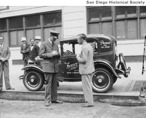 Officer Leo Magone writing a parking ticket for Shrimp Hubbard for parking the Sensor Studio automobile on the sidewalk