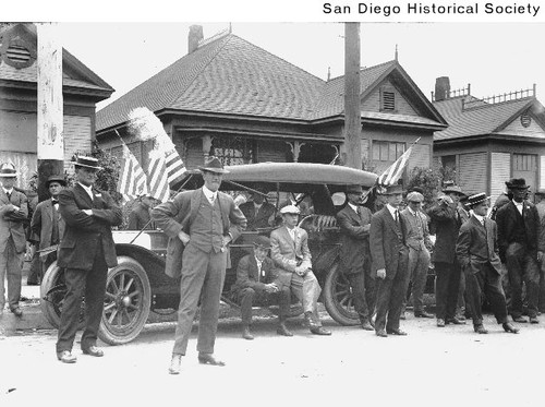 Group of men standing around a flag-draped automobile parked in front of a house