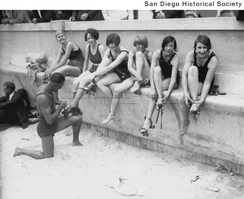 Women in bathing suits putting on roller skates, one receiving assistance from a man in a bathing suit