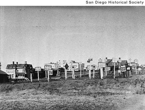 Large number of For Sale signs on a vacant lot with houses in the distance