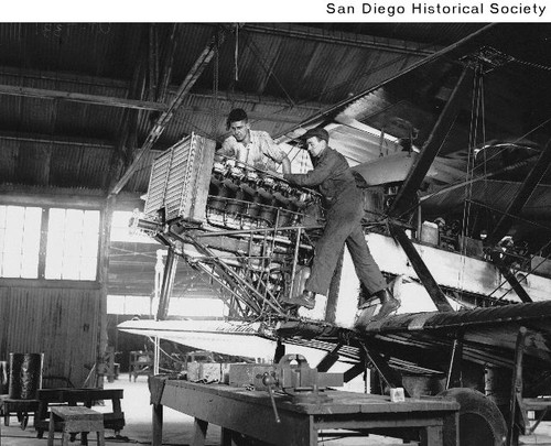 Two men working on the engine of a biplane in a hangar at Rockwell Field