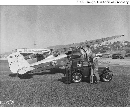 An armed man holding a mail sack and a pilot standing near a U.S. Mail truck parked in front of an airplane
