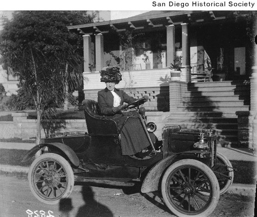 A woman seated at the wheel of an REO Runabout parked in front of a house