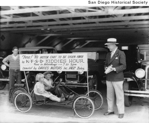 Two children sitting in a motor cart parked in front of a line of automobiles