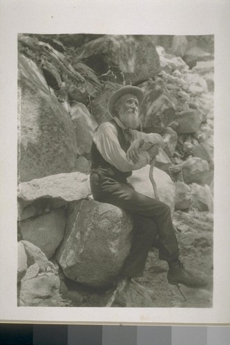 [John Muir sitting on a boulder, with arm resting on staff.]