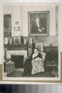 [Gertrude Stein seated in an armchair underneath her portrait by Picasso.]