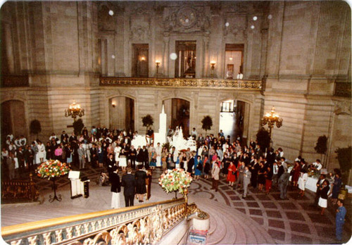[Group of people gathering around a miniature structure of San Francisco while listening to a speaker in the Rotunda of City Hall]