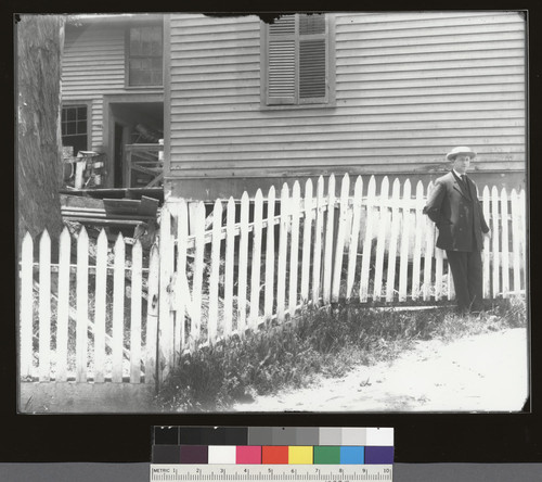 [Man next to fence which drops significantly. (In front of wood-frame building).]