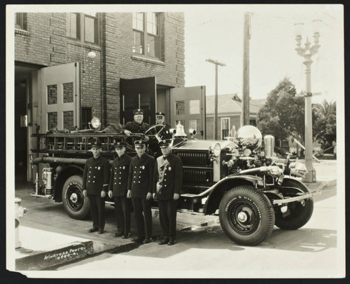 Uniformed firefighters stand in front of ladder truck at Station No. 5, Anaheim & Newport