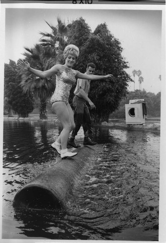 Photo of a woman in a bathing suit, and a lumberjack rolling a log in a pond with an electric dryer on the bank in the background