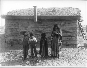 Four Mojave Indian children standing in front of a small dwelling with a thatched roof, near Yuma, ca.1900