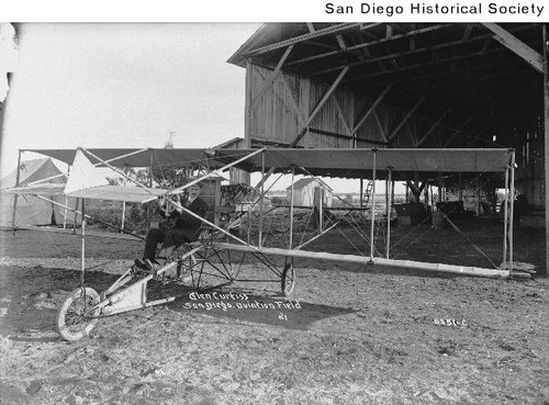 Glenn Curtiss seated in a biplane in front of a hangar on North Island