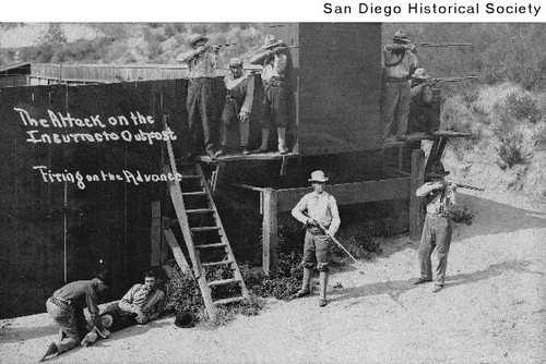 Armed men standing behind a wall with one tending a wounded man