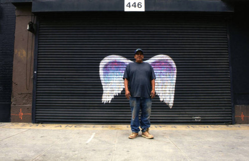 Unidentified man in jeans and baseball cap posing in front of a mural depicting angel wings