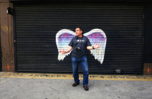 Unidentified man posing with camera in front of a mural depicting angel wings