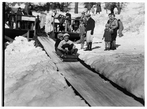 Two boys on a sled riding down a mountain slope while a crowd watches