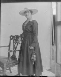 Photo portrait of a woman, standing next to a chair, wearing a long dress, with jewelry and a large hat