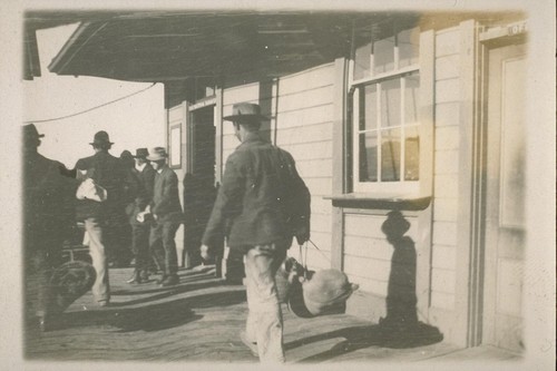 "Blanket Stiff". [Men carrying bedrolls and belongings in front of unidentified building.]