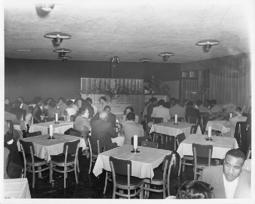Men and women seated at white cloth-covered tables in open dining area of Slim Jenkins Bar and Restaurant Oakland, California
